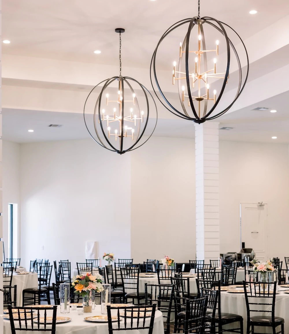 A bright wedding reception ballroom set with round white linen tables, black chiavari chairs, and large modern black and gold orb chandeliers.