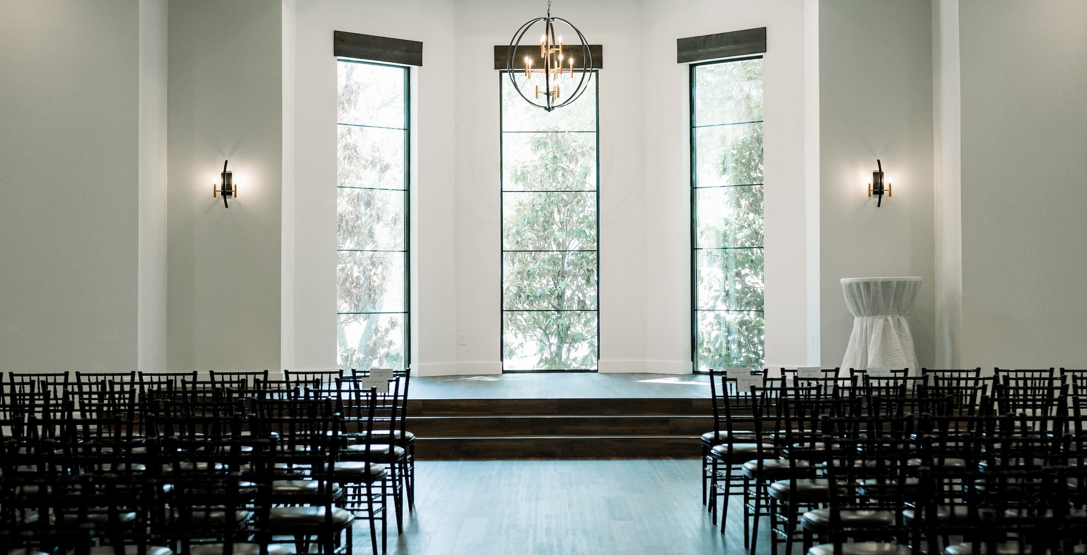 Bright modern wedding ceremony space with floor-to-ceiling windows, black chiavari chairs, and a central chandelier