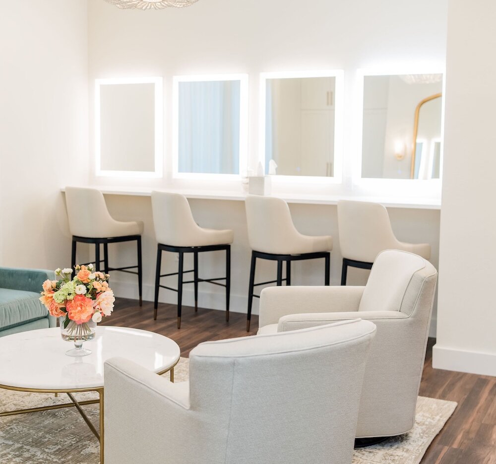 Bright and modern bridal suite vanity area with four illuminated square mirrors and matching light-colored chairs, next to a seating area featuring neutral armchairs, a teal sofa, and a colorful floral centerpiece.
