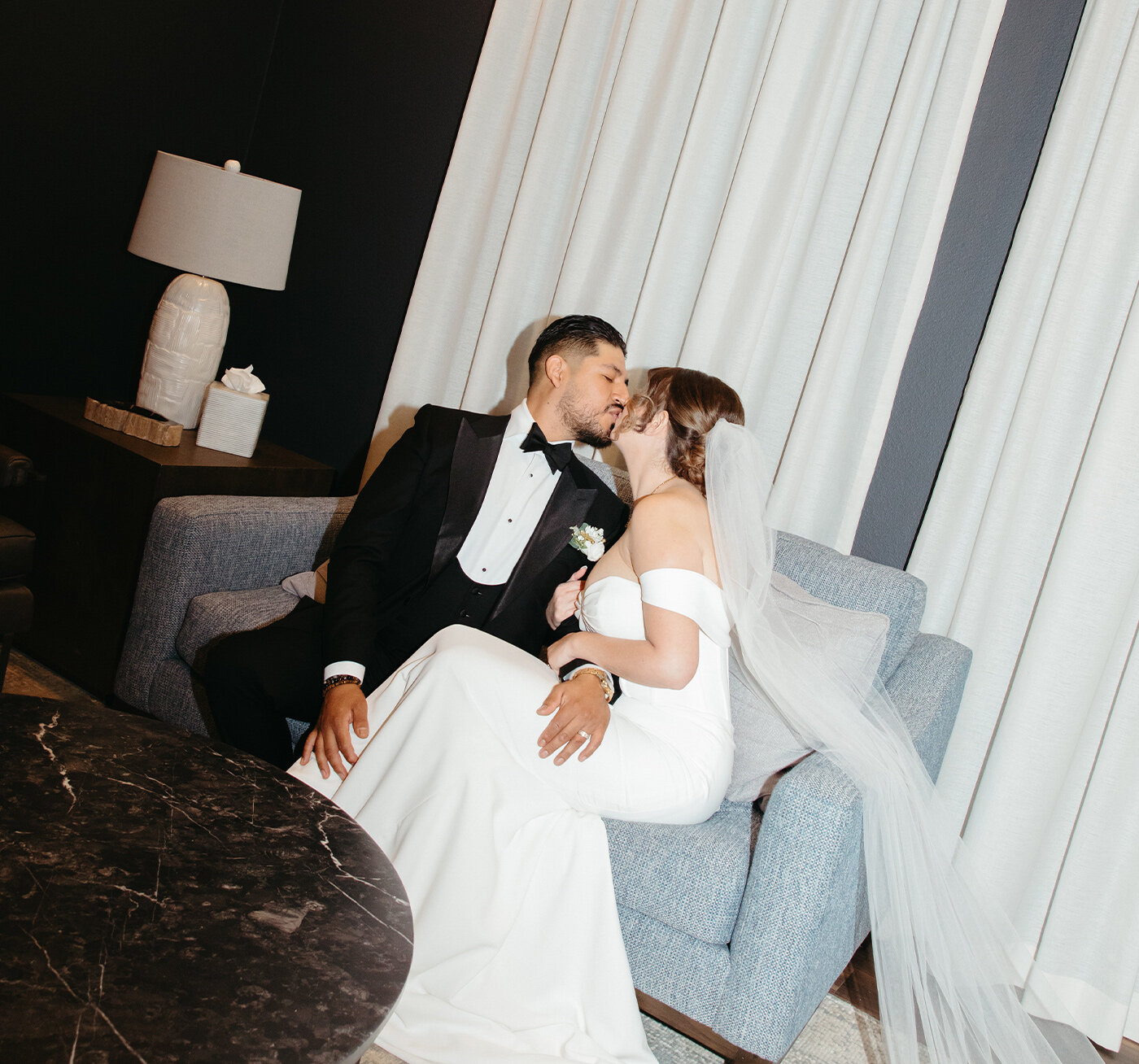 The modern interior of a wedding chapel with dark wood floors, rows of black chiavari chairs, and a sheer draped altar arch set against three large windows.