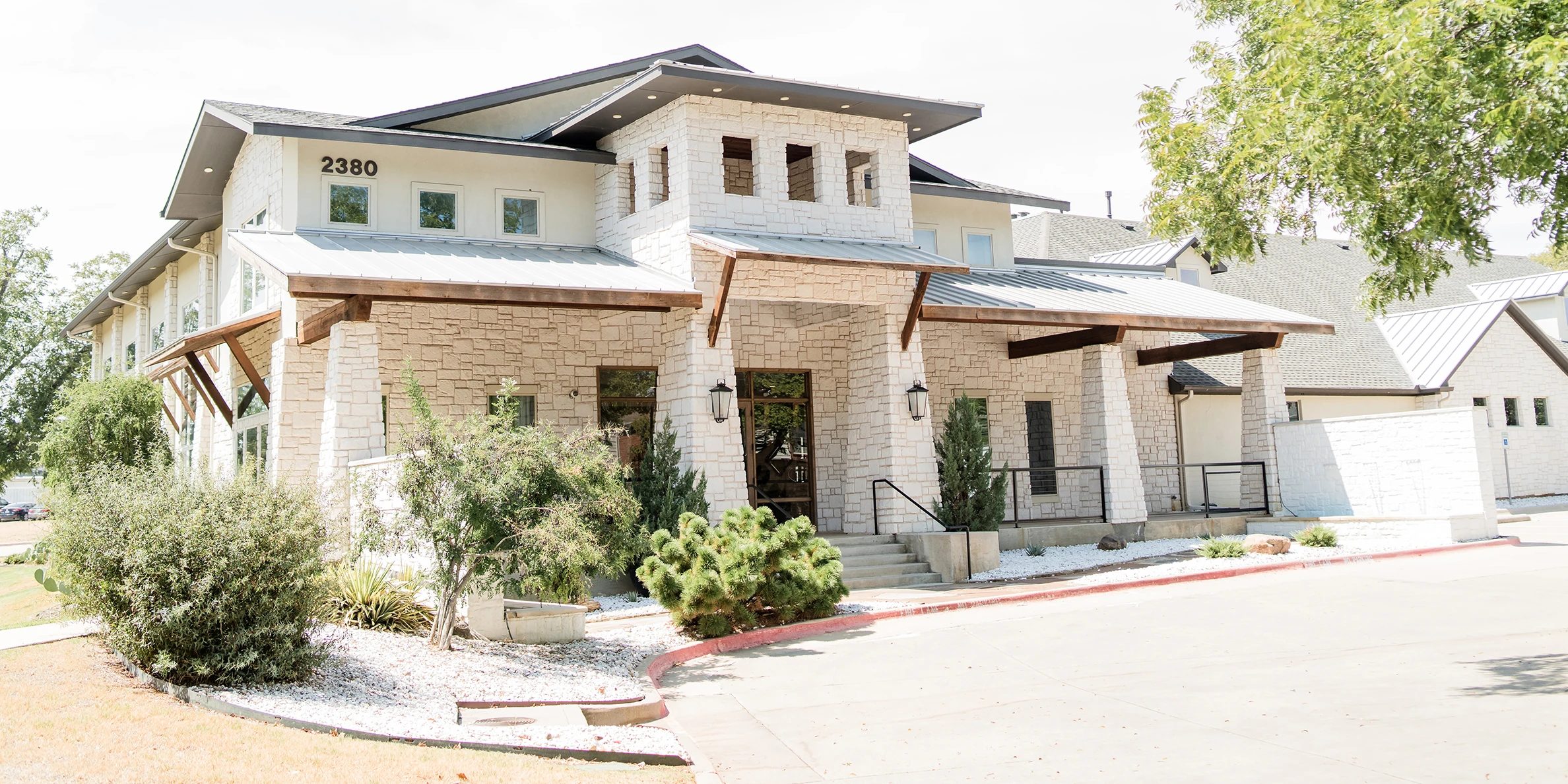 Modern stone building with metal roofing, wooden awnings, and a central tower-style entrance, surrounded by landscaped shrubs and trees on a bright sunny day.