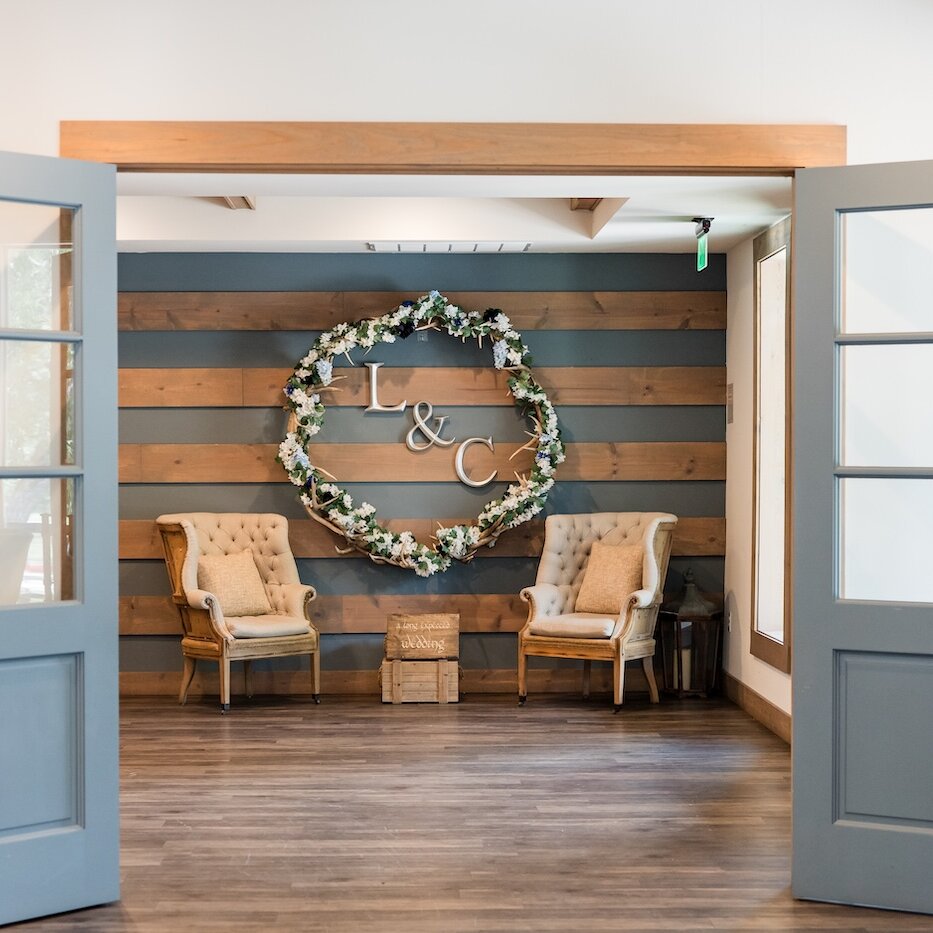 Rustic wedding venue reception entrance with open light blue French doors leading to a lounge area. The focal wall features horizontal wood paneling, two tufted armchairs, and a large wreath with white florals surrounding the initials "L & C".