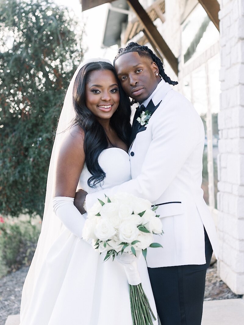 Elegant wedding portrait of a Black couple embracing outdoors in front of a white stone and wood venue entrance. The groom wears a white tuxedo jacket, and the bride wears a strapless white gown with long gloves, holding a cascading bouquet of white roses.