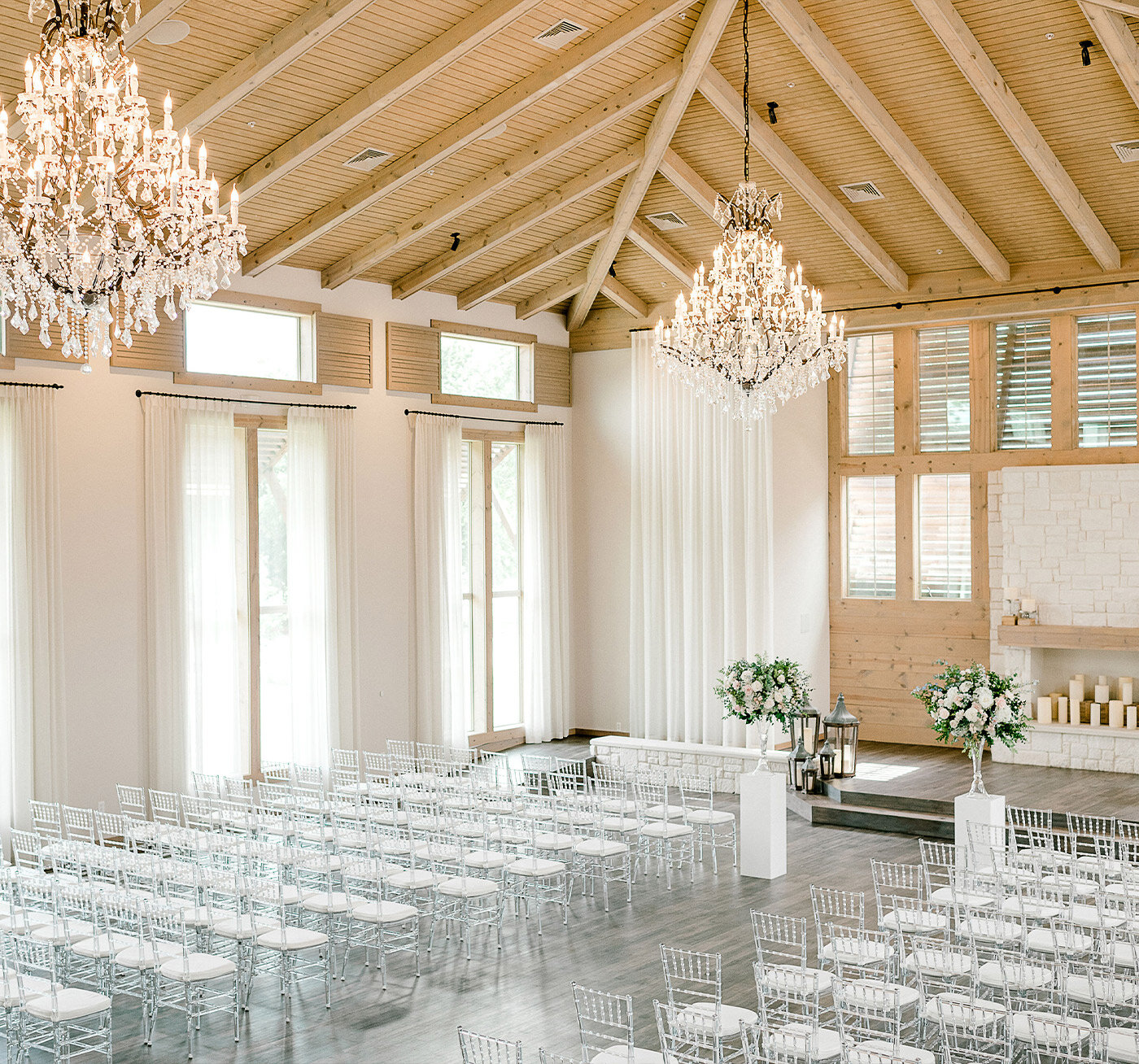 Indoor wedding ceremony setup with a stone fireplace decorated with floral arrangements in pastel colors and rows of clear chairs