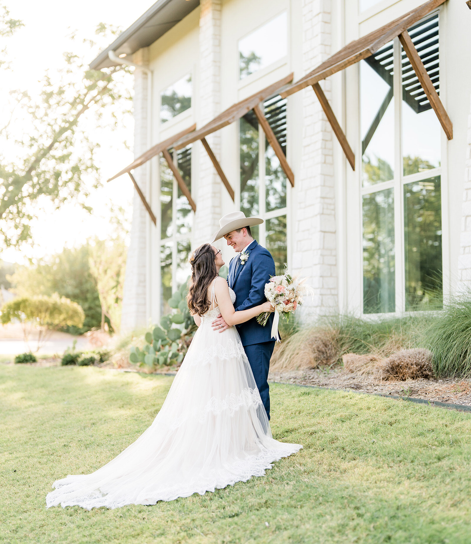 Stylish wedding portrait of the groom and an elder man posing indoors in black-tie attire and sunglasses. The groom wears a white tuxedo jacket, and the man next to him wears a traditional black tuxedo, standing in a bright venue with exposed wood beams and white curtains.