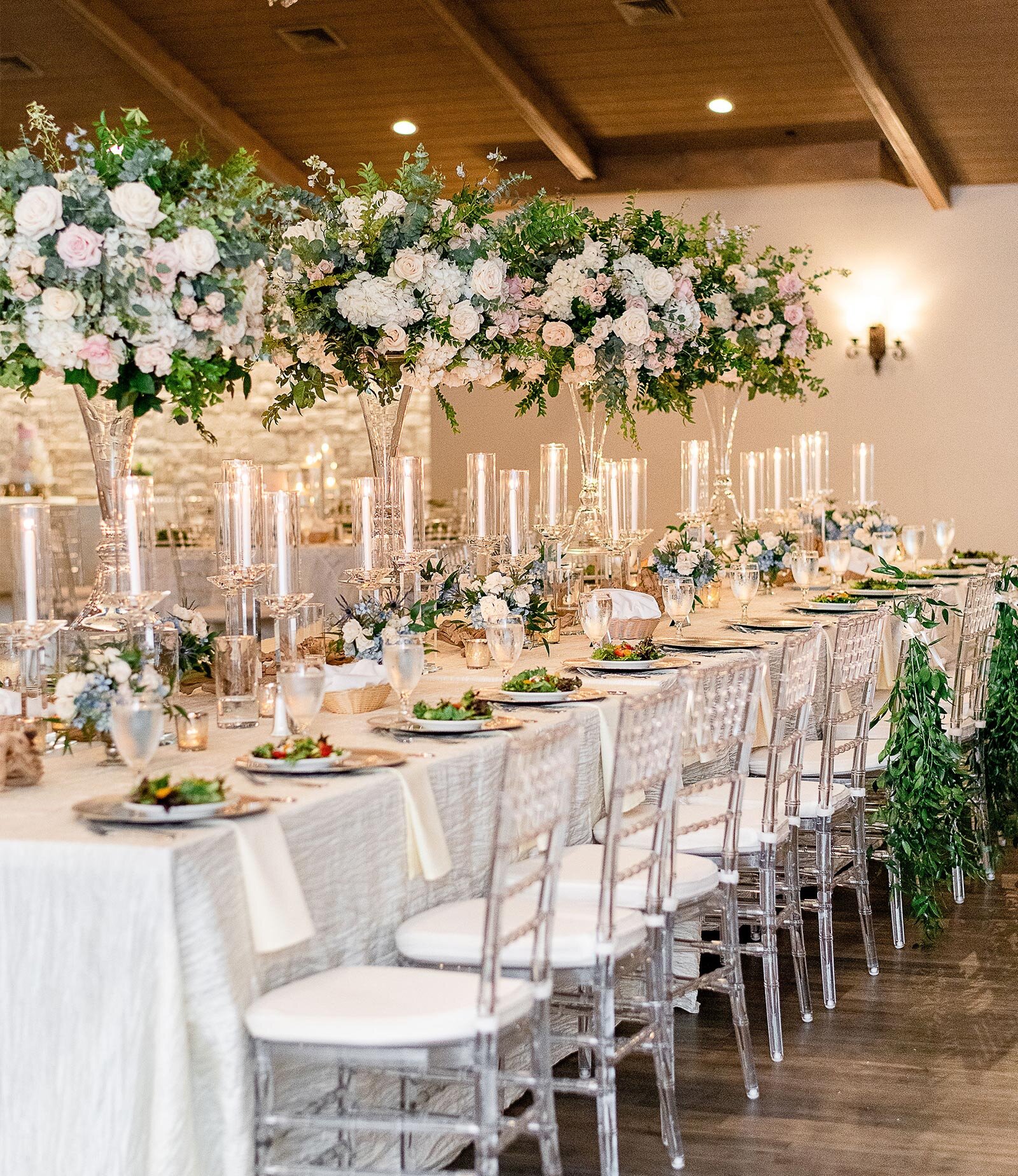 Modern wedding venue exterior featuring stacked light-colored limestone facade and angular architecture with a prominent stone entryway. The entrance has large glass doors, wood beams, and is framed by juniper trees and white rock landscaping.