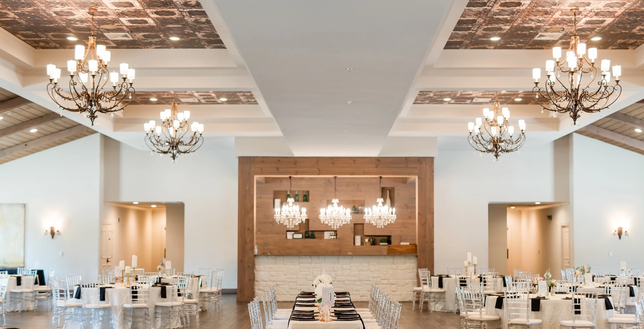 A large wedding reception ballroom featuring a white and copper pressed tin ceiling, chandeliers, and a central bar area with light wood paneling.