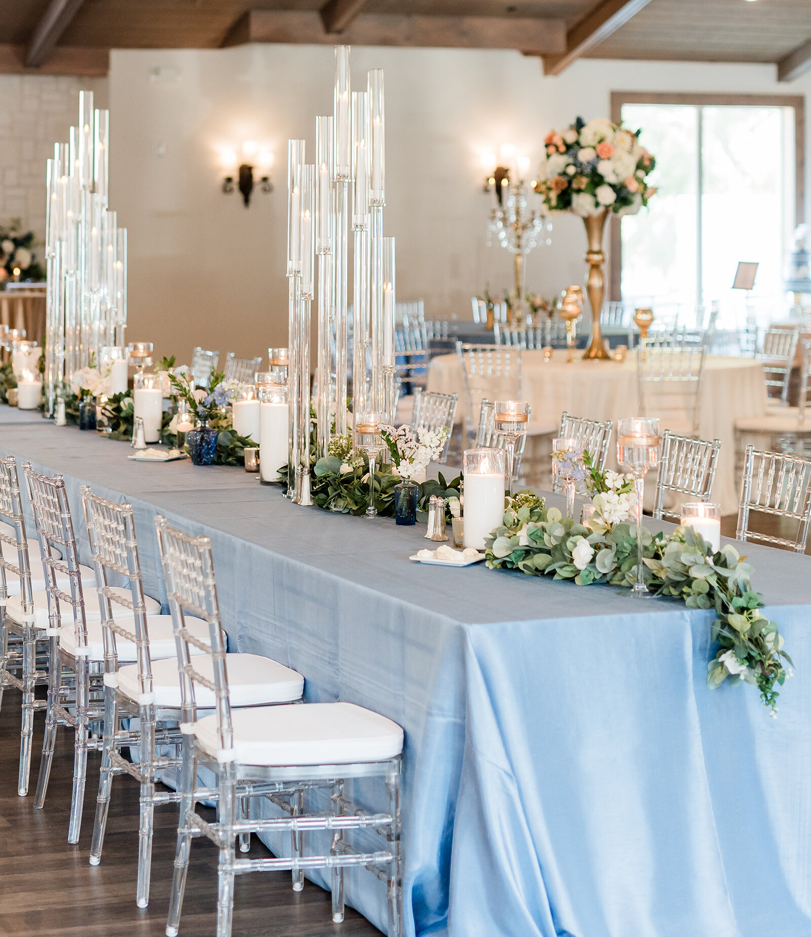 Elegant wedding reception table with light blue linens, clear acrylic chairs, tall glass candle centerpieces, and greenery floral garlands