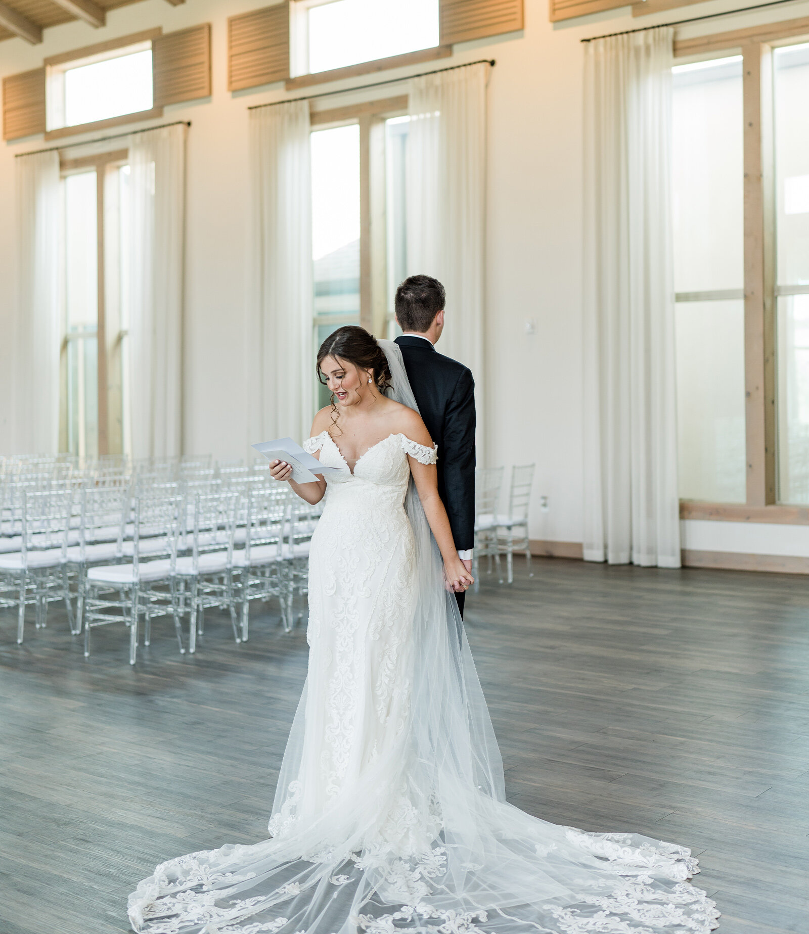 The bright interior of a wedding ceremony hall featuring a vaulted wood ceiling, crystal chandeliers, a brick fireplace, and rows of clear acrylic chairs.