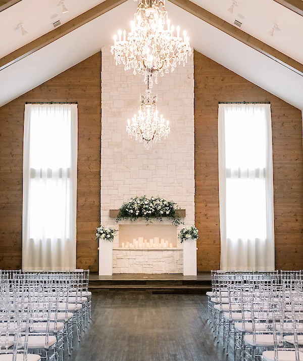 Modern wedding venue exterior entrance featuring white stacked stone, stucco, and wood accents under a metal awning. The entrance has dark double doors and is framed by lush trees, including one with pink blossoms.