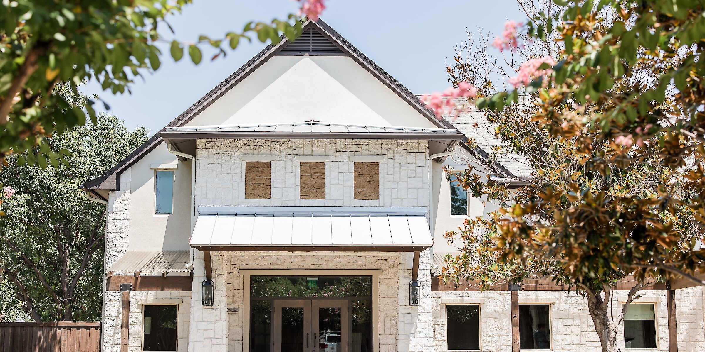 Modern wedding venue exterior entrance featuring white stacked stone, stucco, and wood accents under a metal awning. The entrance has dark double doors and is framed by lush trees, including one with pink blossoms.