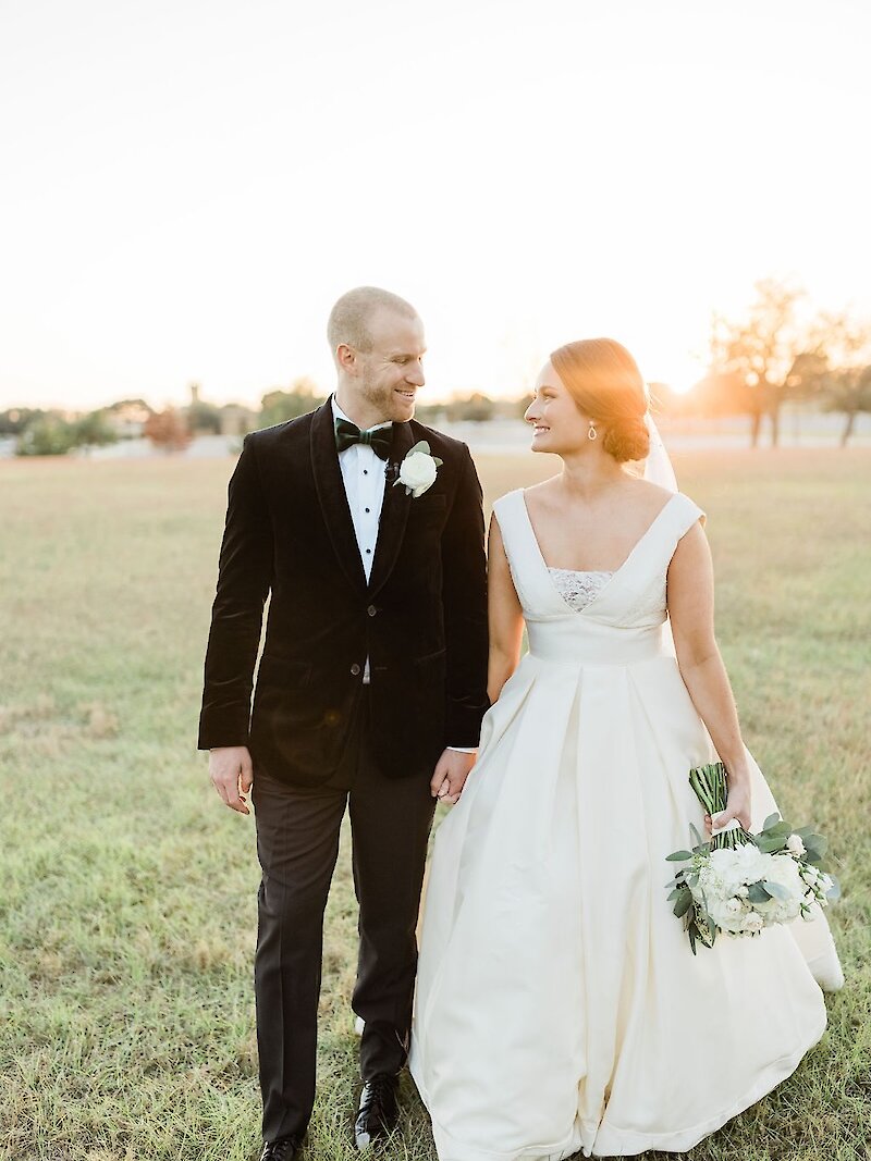 Golden hour wedding portrait of the couple walking hand-in-hand in a grassy field during sunset. The groom wears a black velvet tuxedo jacket, and the bride wears a cap-sleeve satin A-line gown and holds a white hydrangea bouquet.