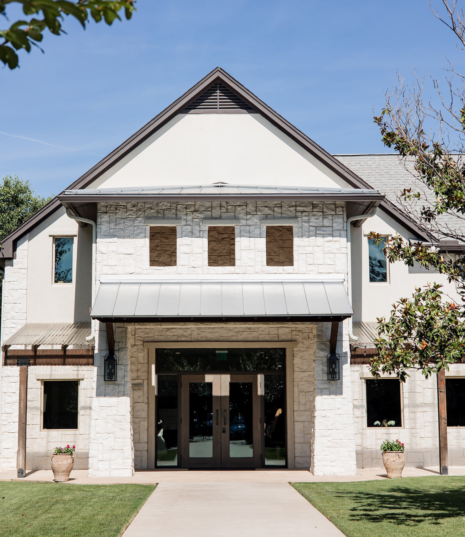 Front entrance of a modern stone and stucco event venue with double glass doors, metal awning, and landscaped walkway on a sunny day