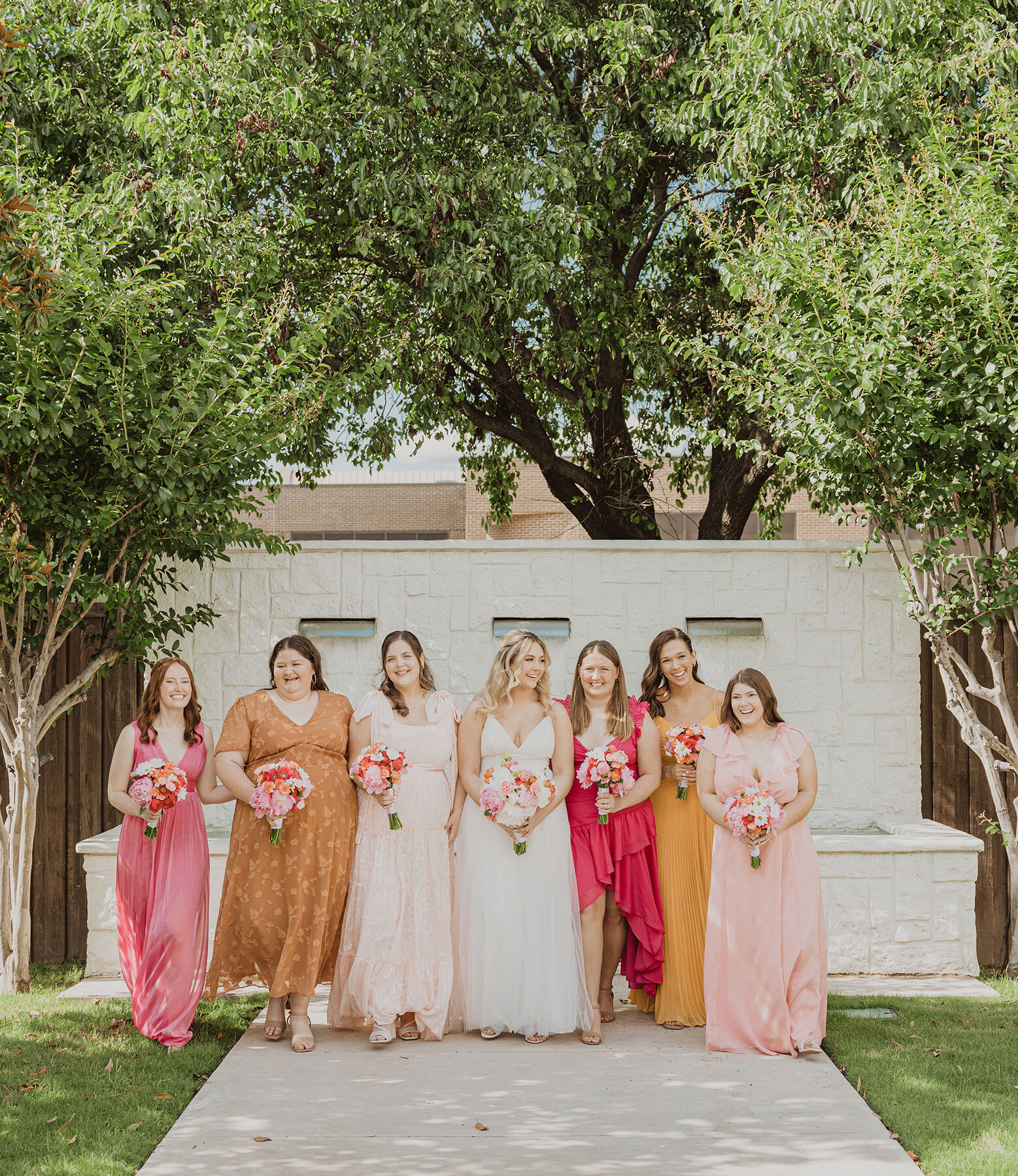 Bride standing with bridesmaids in colorful dresses holding bright bouquets in front of white stone water feature and lush green trees