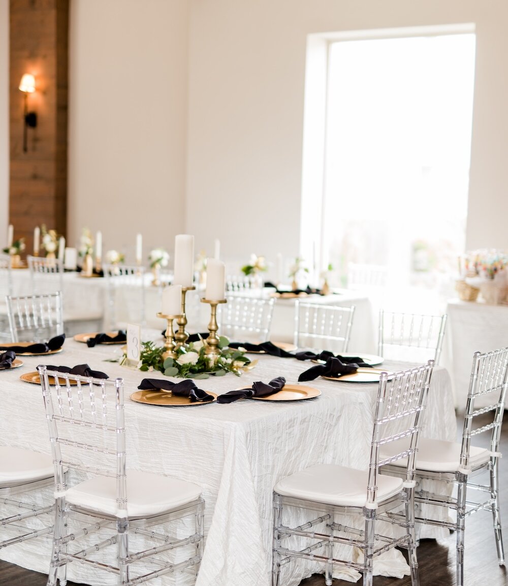 Bright wedding reception table setting with white textured linens, clear ghost chairs, and gold charger plates with black napkins. The centerpiece features tall white candles on gold candelabras and green and white floral accents in a modern, light-filled venue.