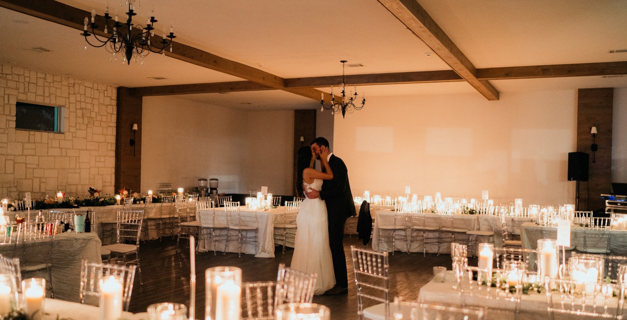 The bride and groom share their first dance in the center of a warmly lit wedding reception room filled with dozens of candles and long dining tables.