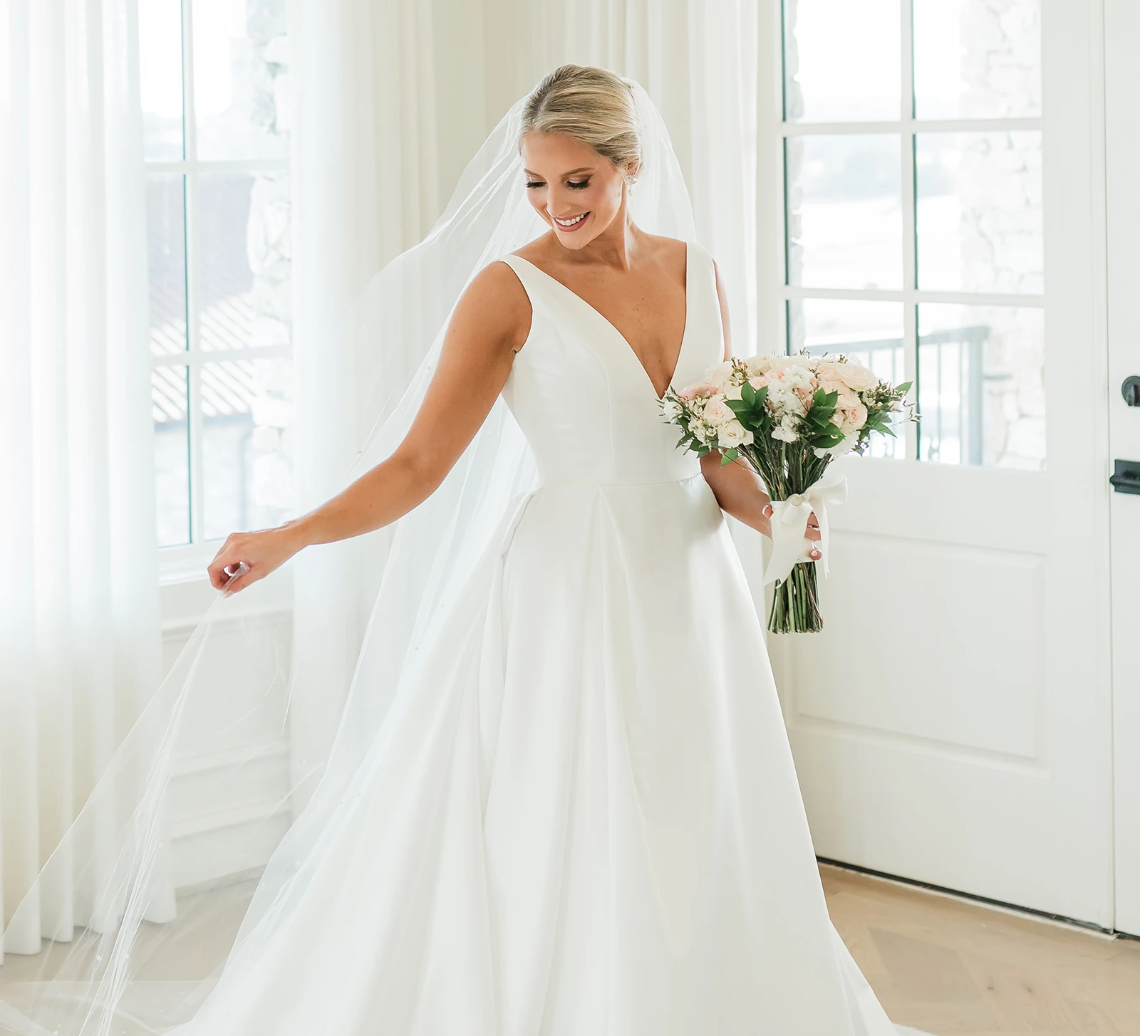 Bride smiling and holding a bouquet while posing in a classic white wedding dress with a long veil in a bright, natural-light room