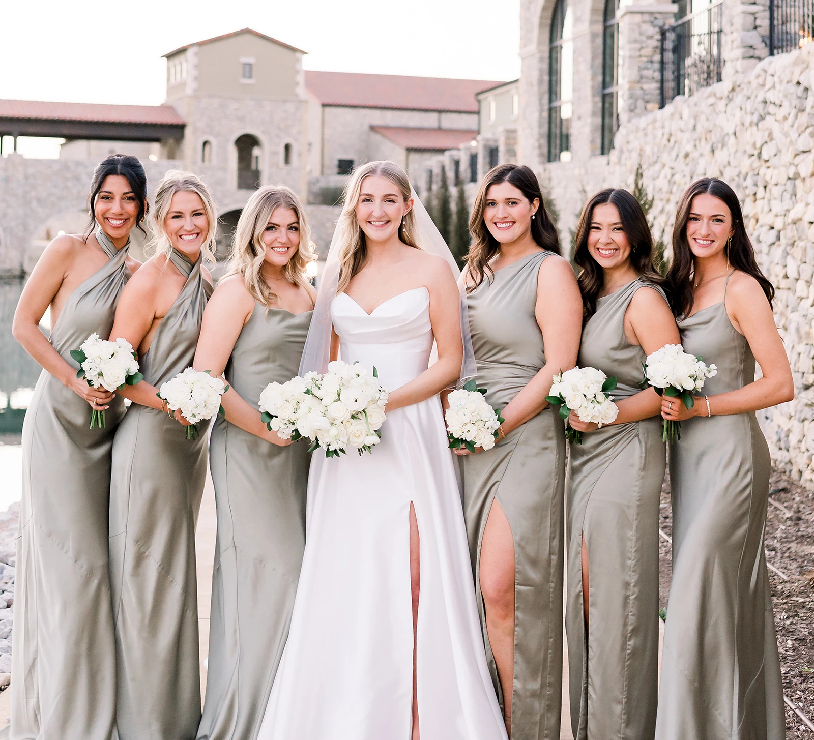 Bride standing with bridesmaids in sage green dresses holding white bouquets at a waterfront stone wedding venue