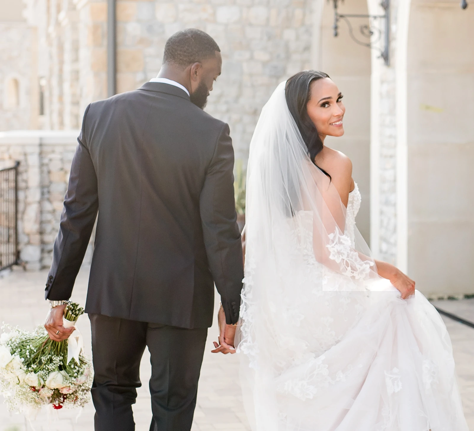 Bride and groom walking hand in hand at a stone wedding venue, with the bride smiling back in a lace gown and veil
