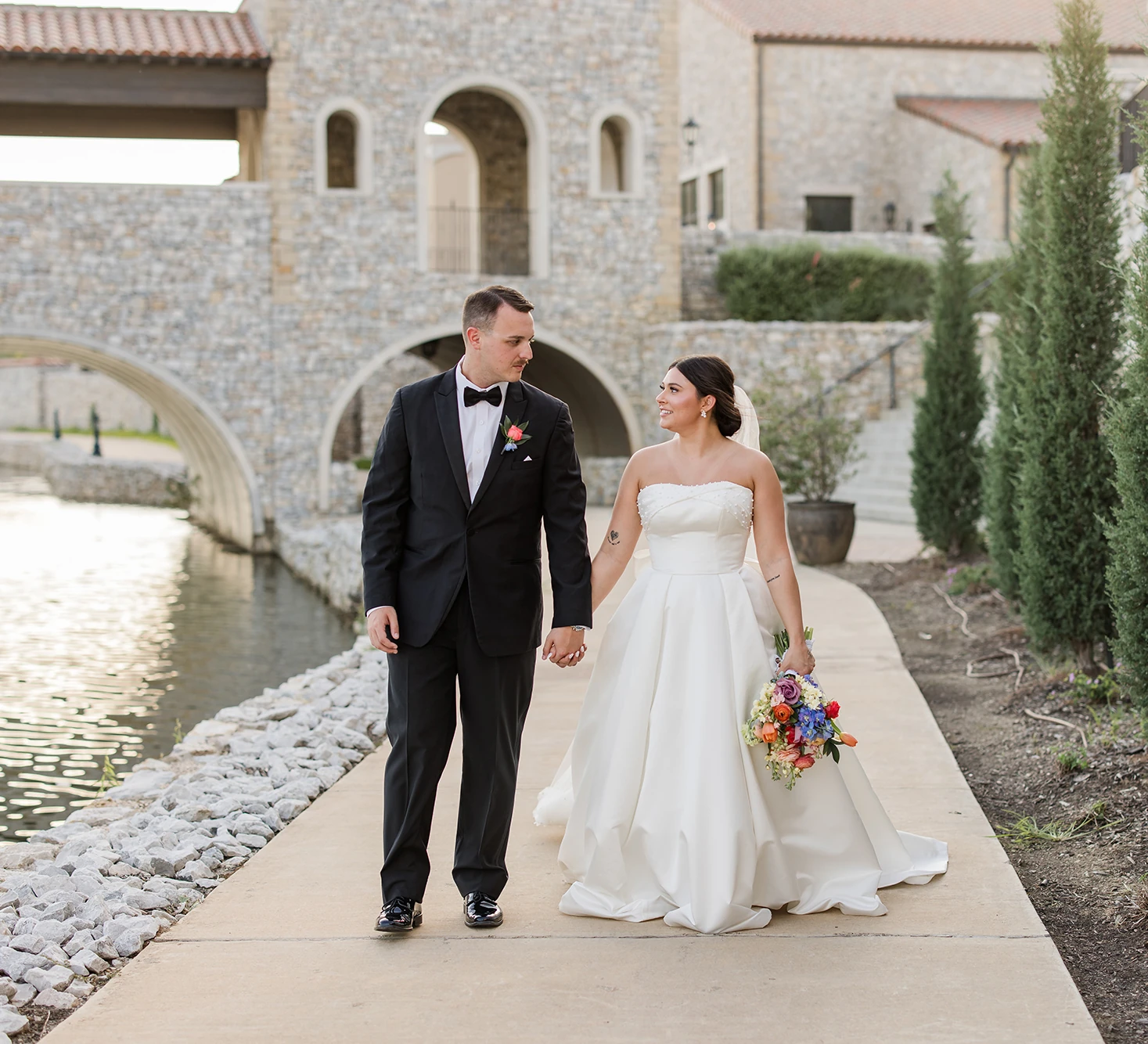 Bride and groom walking hand in hand along a waterfront path with a stone bridge and Italian-inspired wedding venue in the background