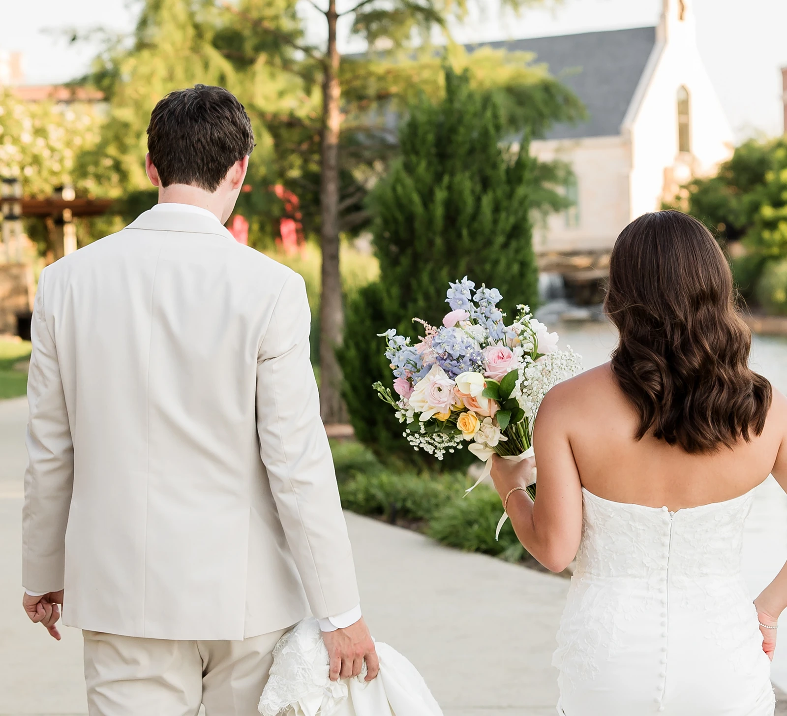 Bride and groom walking away together outdoors, with the bride holding a pastel floral bouquet and wearing a strapless gown