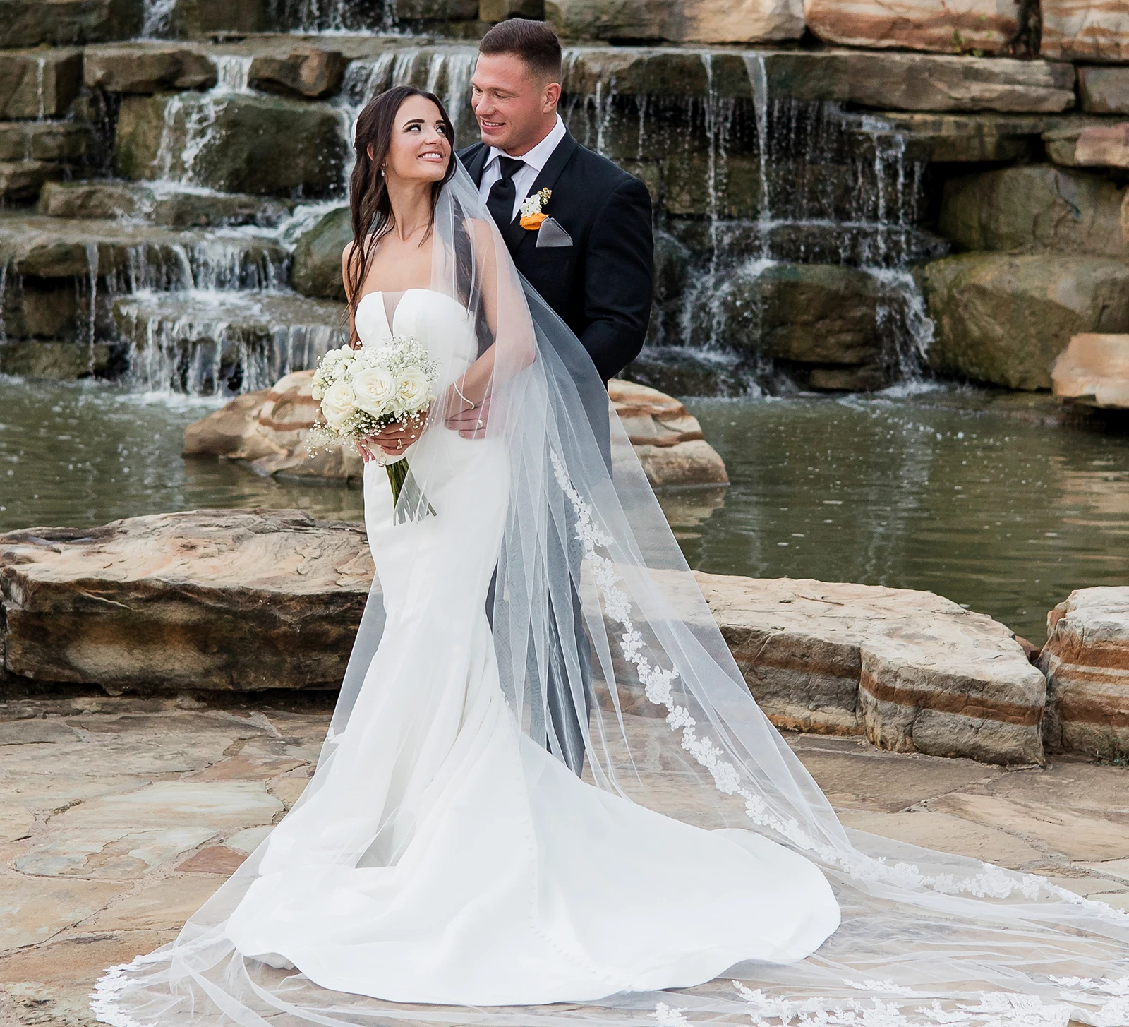 Bride and groom posing in front of a waterfall, with the bride’s long veil flowing and the groom smiling at her.