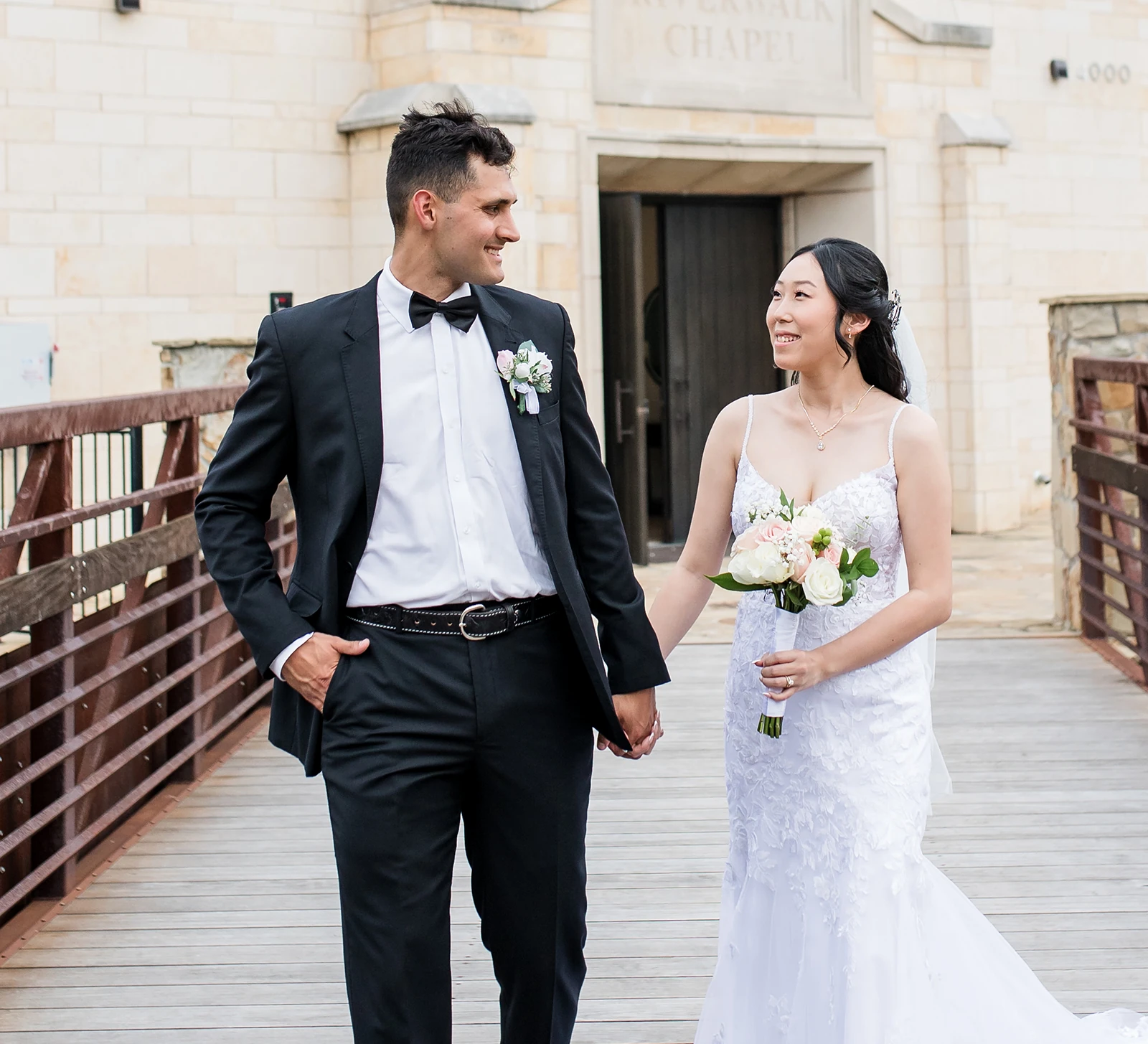 Bride and groom walking hand in hand across a wooden bridge outside the Riverwalk Chapel, smiling at each other on their wedding day