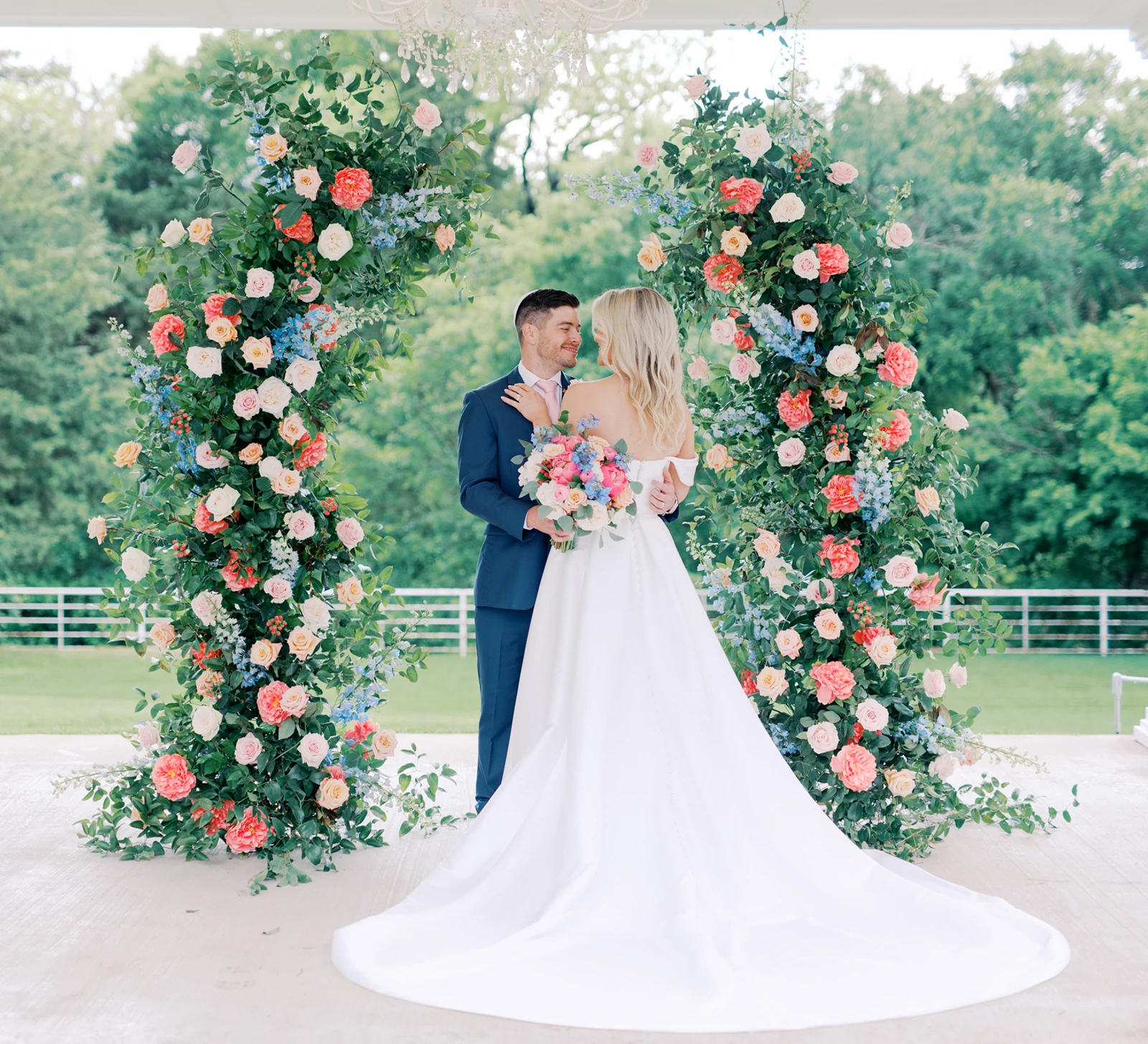 Bride and groom standing under a vibrant floral arch during their outdoor wedding, with the bride in an off-the-shoulder gown and long train holding a colorful bouquet