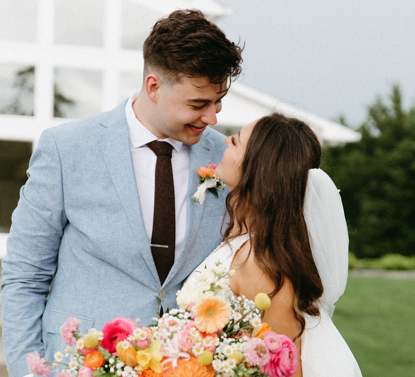 Bride and groom smiling closely at each other outdoors, with the bride holding a colorful spring bouquet of pink, orange, and yellow flowers