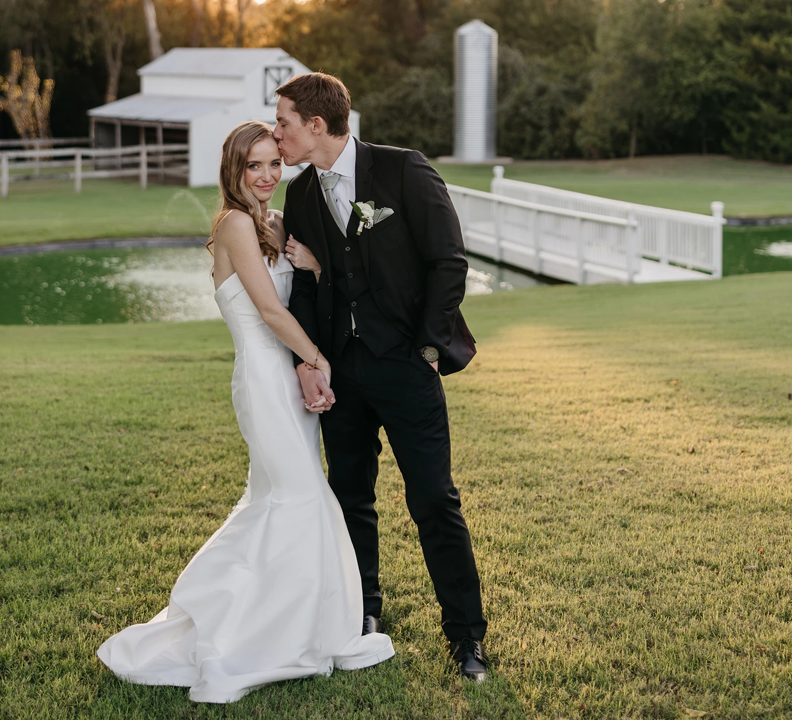 Groom kisses bride on the forehead during sunset at an outdoor wedding, standing on a lawn near a pond and white bridge