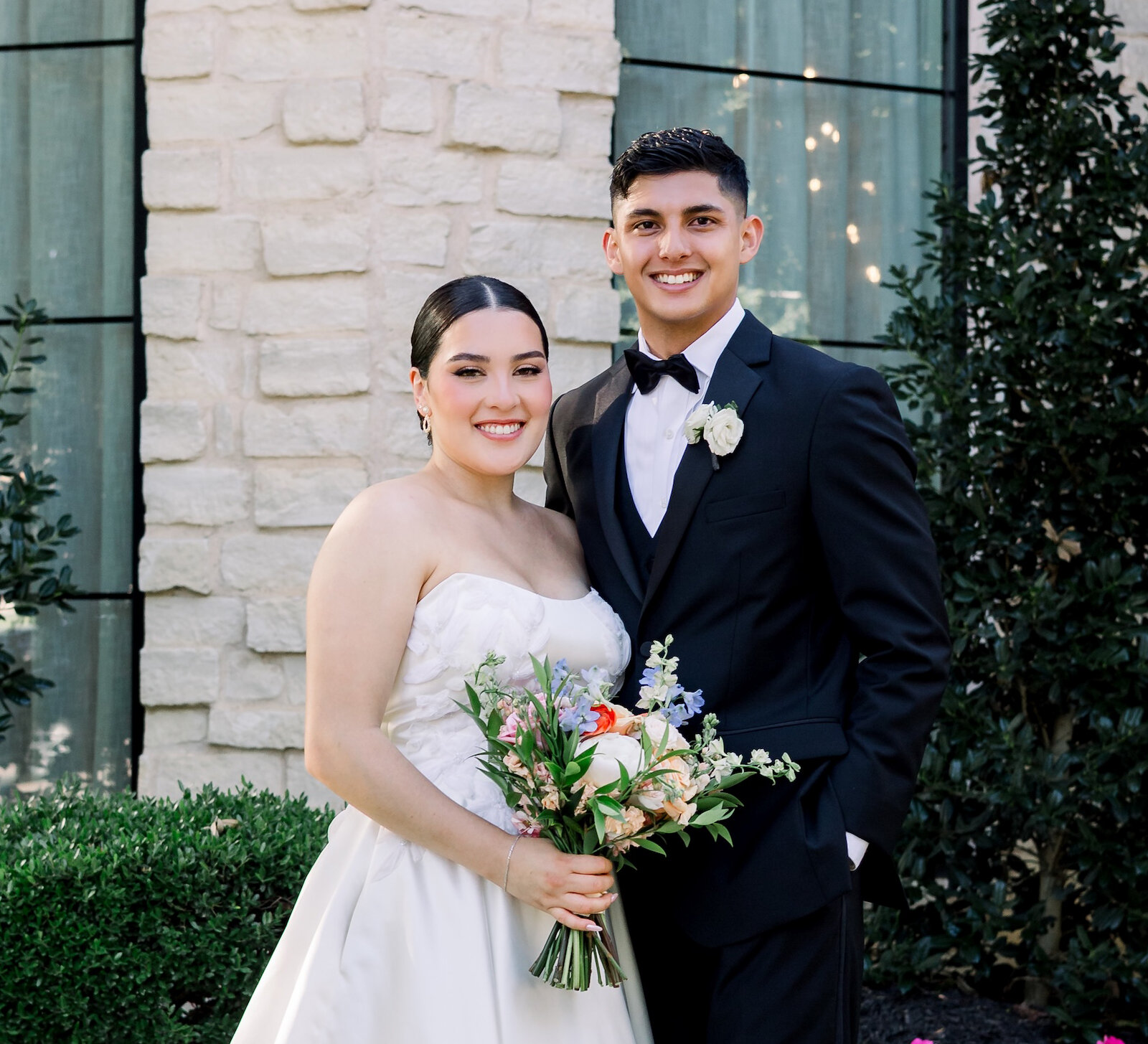 Bride and groom smiling in an outdoor wedding portrait, bride in a strapless white gown holding a pastel bouquet, groom in a black tuxedo