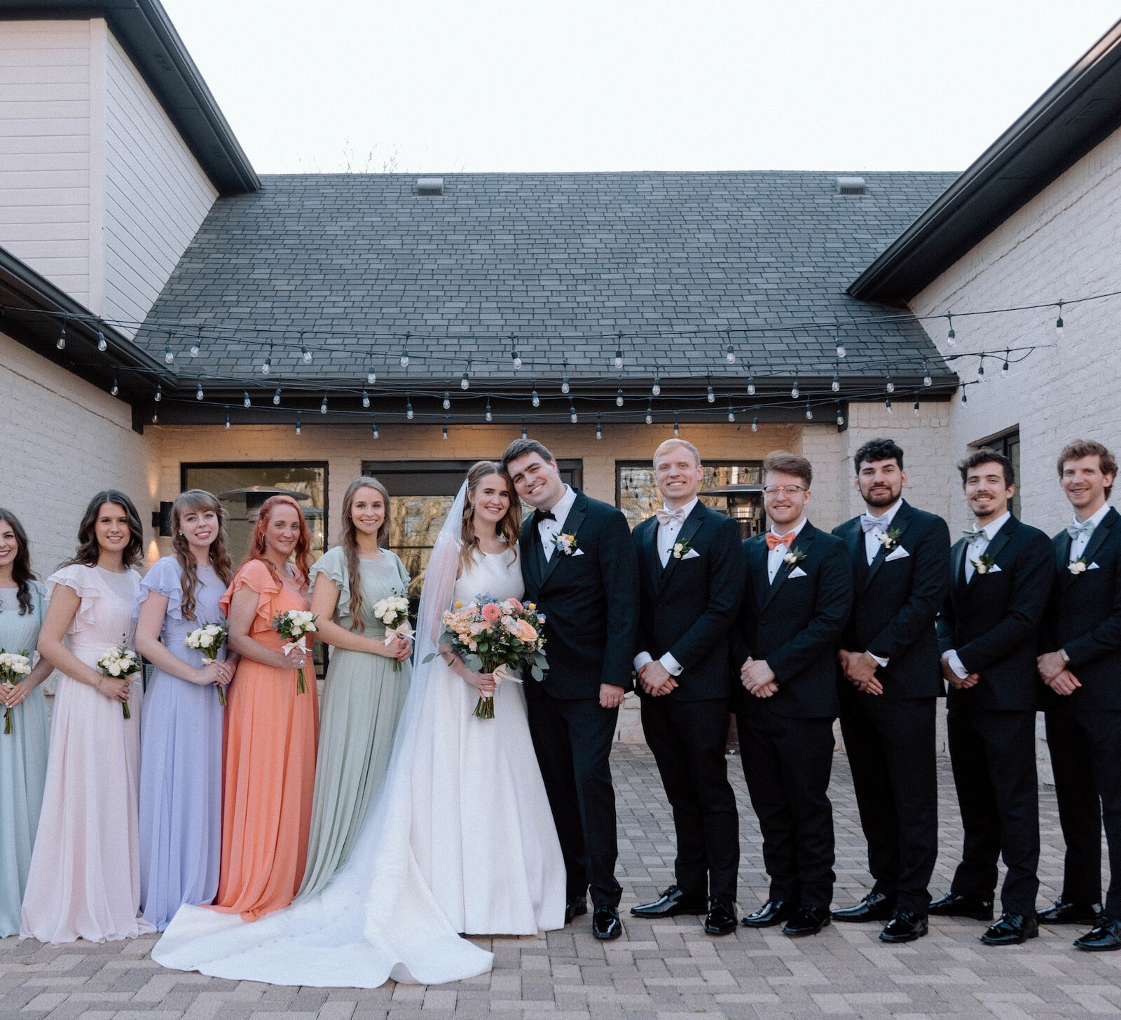 Wedding party portrait outside a modern venue with a black roof and white stone facade. The bride and groom are surrounded by bridesmaids in mismatched pastel gowns (blue, pink, lavender, sage, and coral) and groomsmen in black tuxedos.