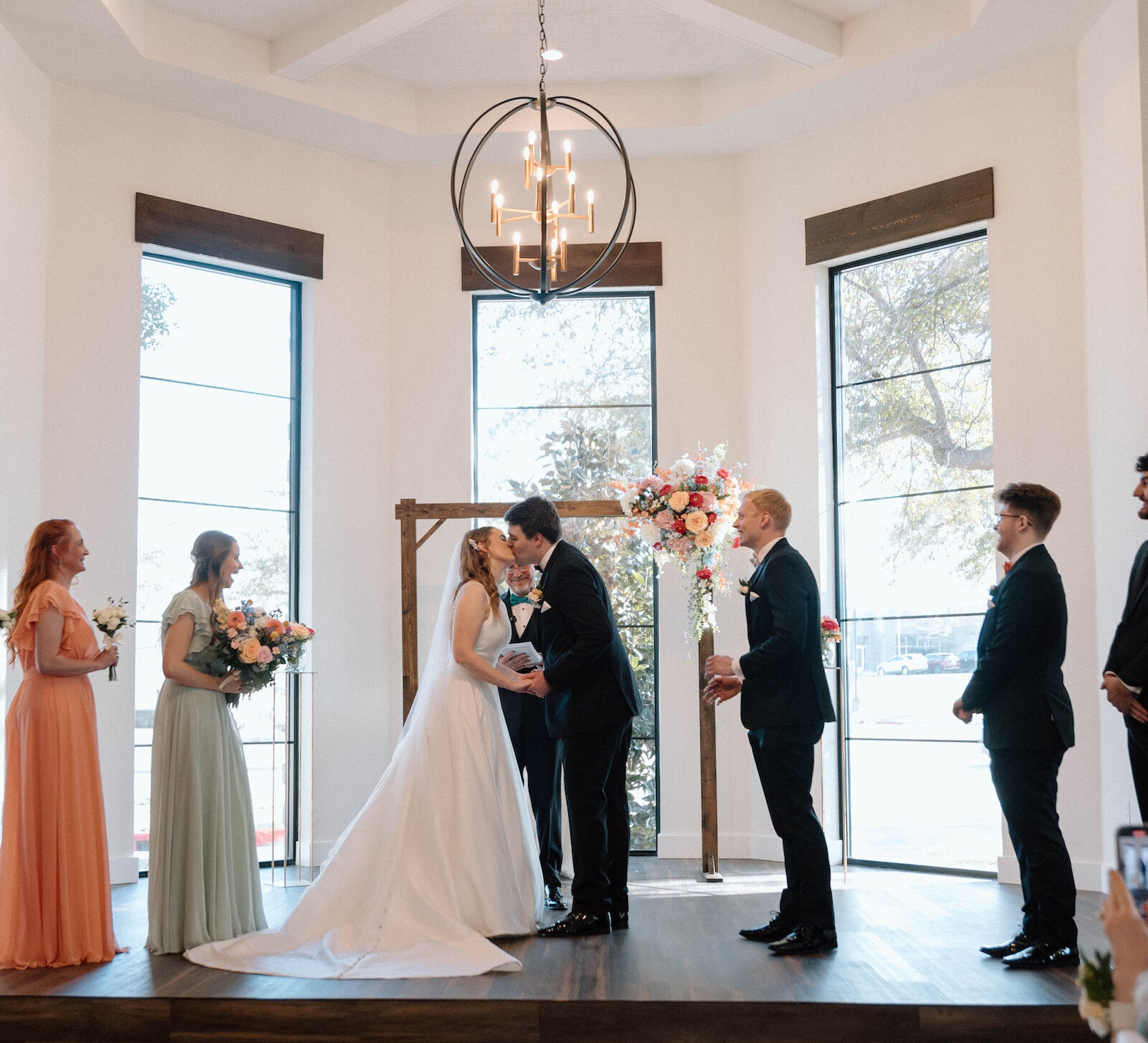 Intimate wedding ceremony kiss inside a brightly lit modern chapel with large vertical windows and a circular iron chandelier. The couple stands under a wooden arch, flanked by the wedding party in mismatched pastel gowns and black tuxedos.