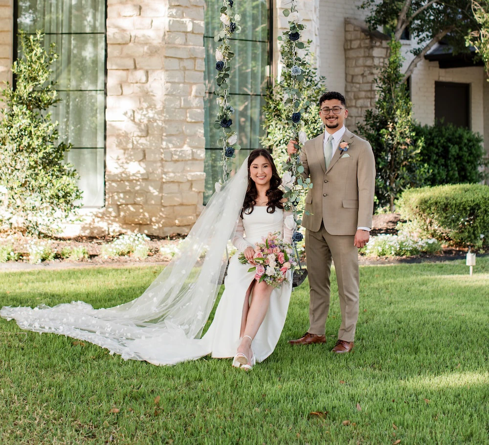 Romantic wedding portrait of an Asian couple standing and sitting on a floral-decorated swing hanging from a large tree. The groom wears a light tan suit, and the bride wears a long-sleeved satin gown with a sweeping veil, set against a light stone venue exterior.