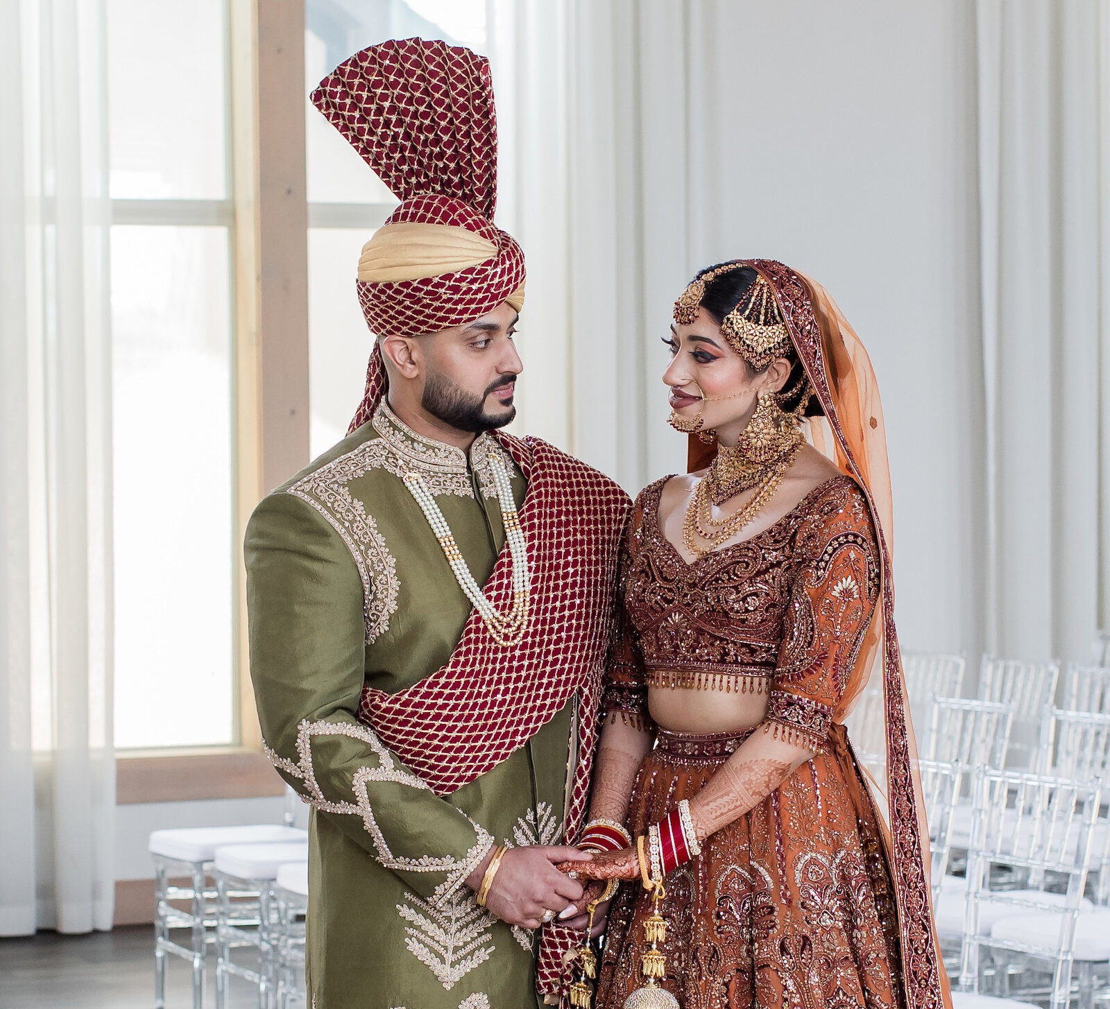 Elegant portrait of a couple in traditional Indian wedding attire standing in a bright, modern venue with large windows. The groom wears an olive green sherwani with a red turban and sash, and the bride wears a heavily embroidered burnt orange and gold lehenga with elaborate bridal jewelry.