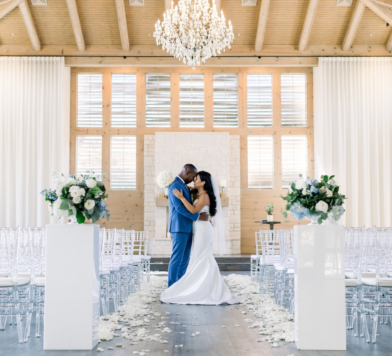 Elegant wedding ceremony photo of the couple kissing at the altar in a bright, modern chapel with exposed wooden beams and a stone fireplace backdrop. The room features a cascading chandelier, white sheer curtains, and rows of clear ghost chairs lining the petal-covered aisle.