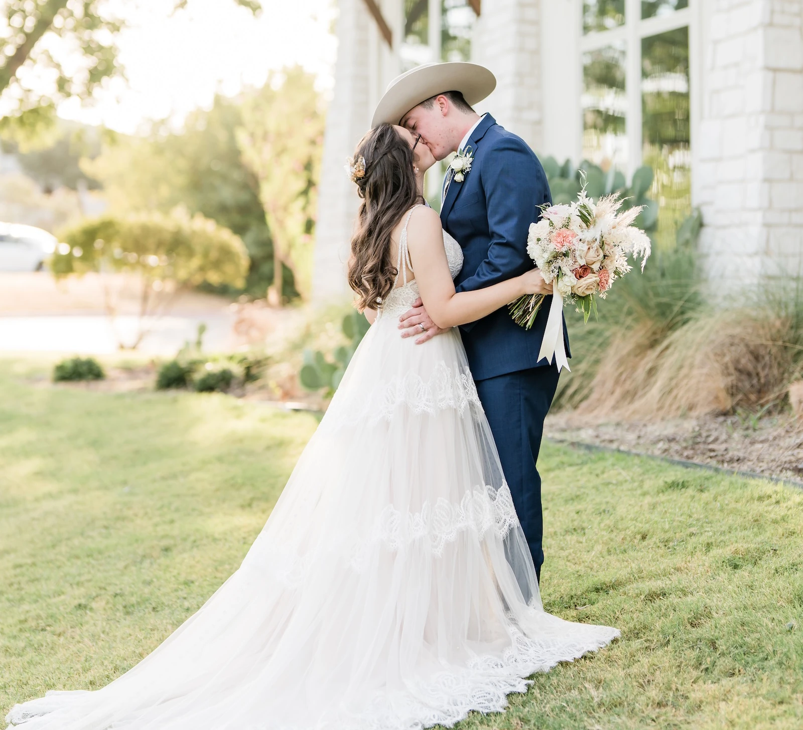 Sunlit wedding portrait of the couple kissing on a green lawn outside a modern white stone venue. The groom wears a blue suit and a cowboy hat, and the bride wears a flowing lace and tulle gown, holding a bouquet with dried grasses and peach flowers.