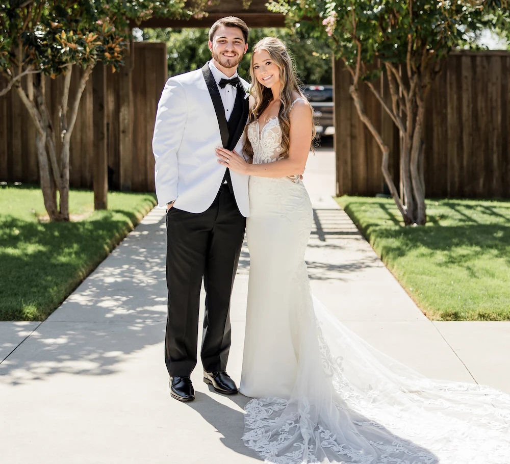 Sunny wedding portrait of the couple posing on a walkway flanked by blooming pink-flowered trees and a wooden fence. The groom wears a white tuxedo jacket, and the bride wears a fitted lace gown with a plunging neckline and long train.