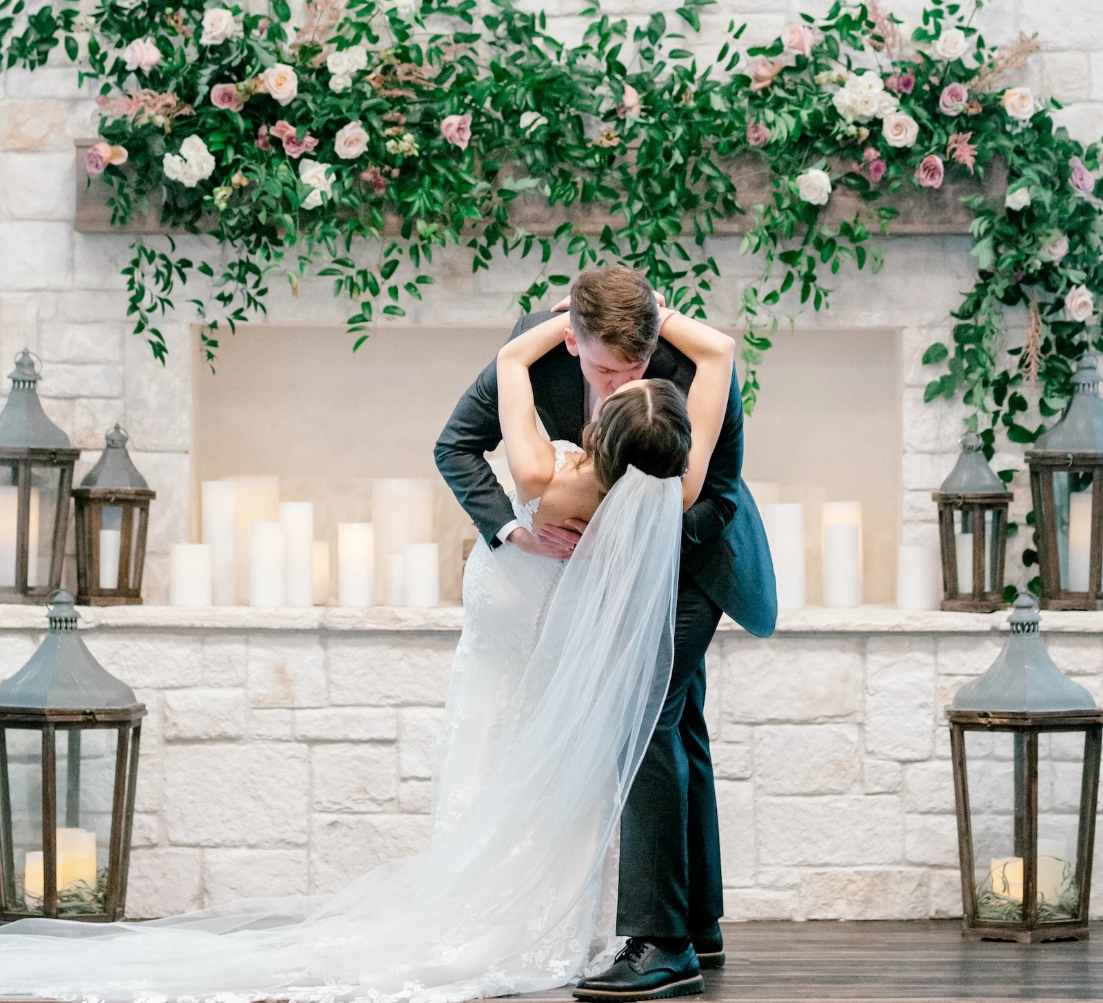Romantic wedding kiss and dip at the ceremony altar, set against a rustic stone fireplace decorated with a lush floral and greenery garland. Large lanterns and pillar candles surround the couple in this indoor barn venue.