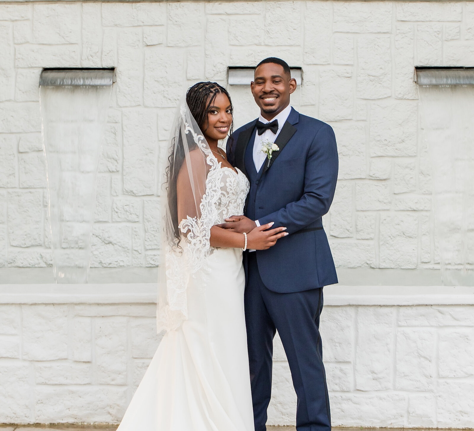 Elegant wedding portrait of a Black couple embracing in front of a modern white stacked stone wall with waterfall features. The groom wears a navy blue tuxedo, and the bride wears a flowing lace gown with a dramatic train and fingertip veil.