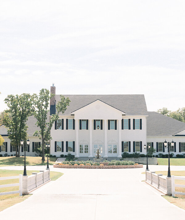Elegant white Southern-style mansion with tall columns, black shutters, and a tiered fountain in front, surrounded by blooming landscaping under a clear blue sky.”