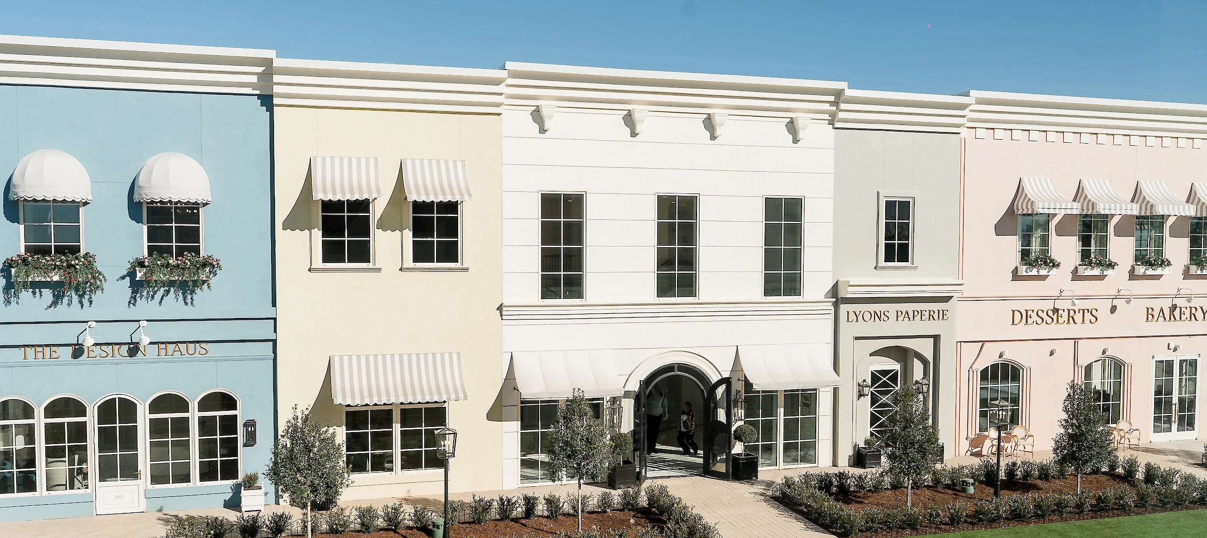 The exterior facade of The Shops at Wedding Square, featuring a row of shops painted in various pastel colors under a clear blue sky.