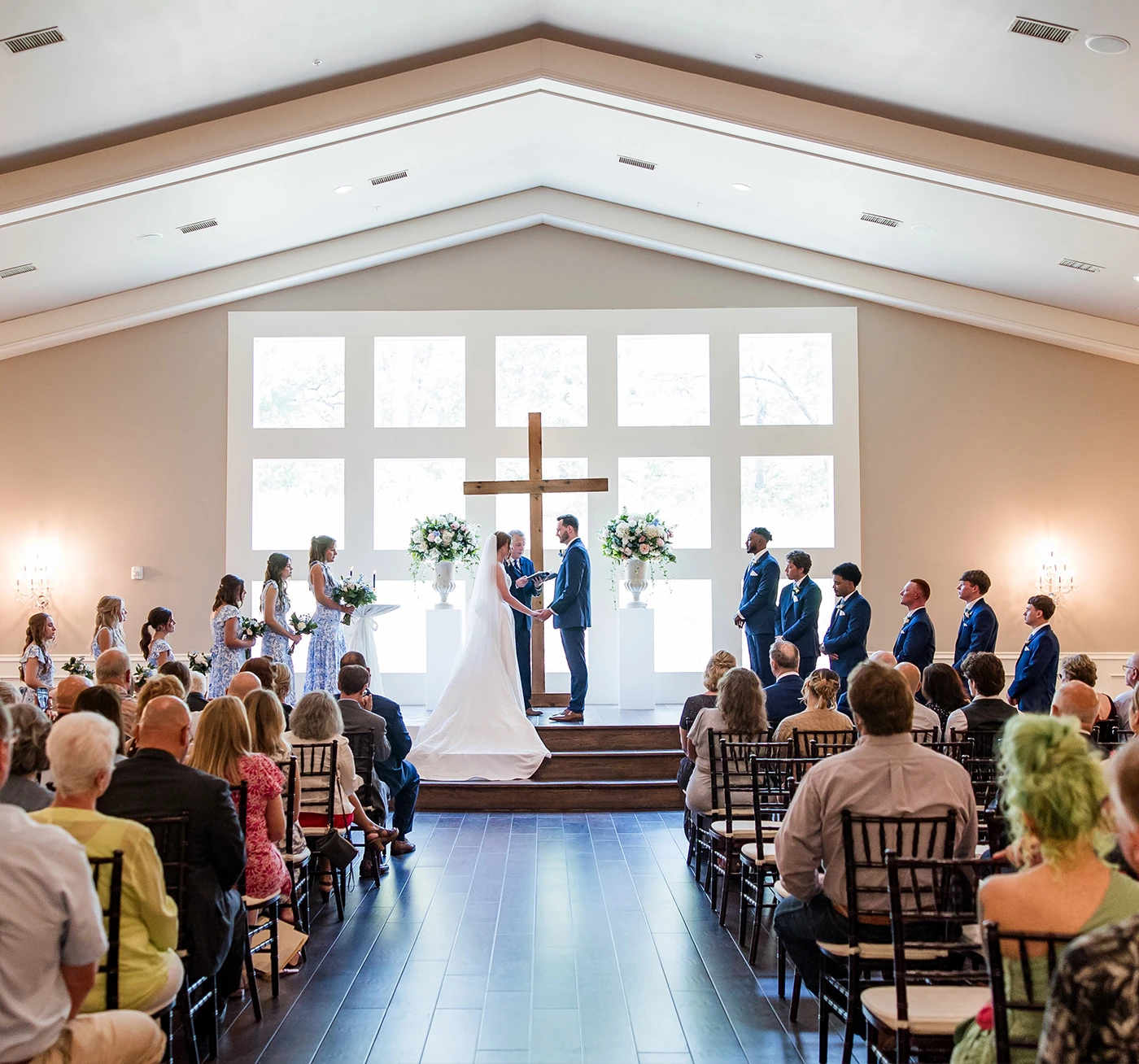 The bride and groom stand closely on a wooden altar, surrounded by light blue and white floral arrangements, in front of tall chapel windows.