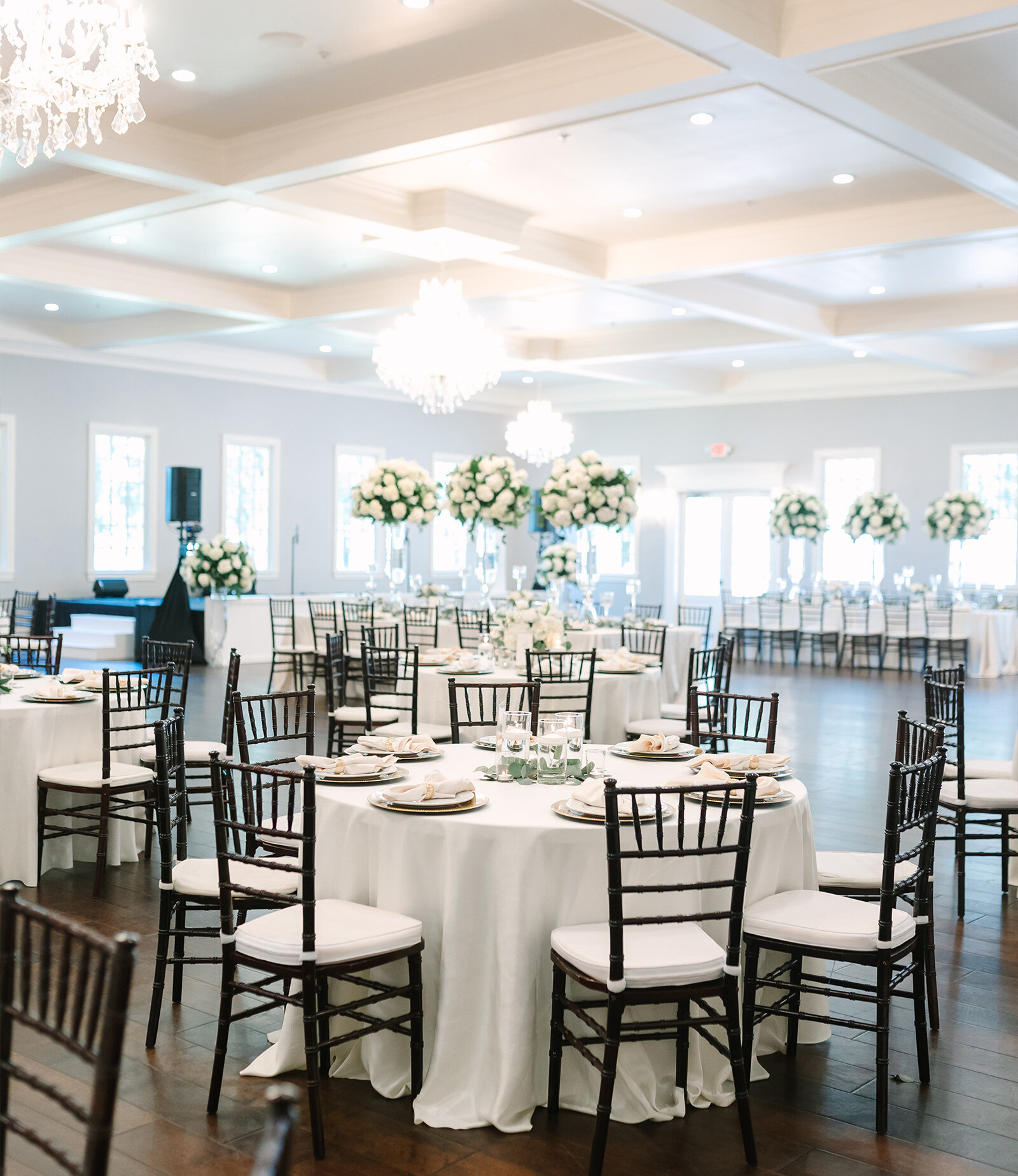 Elegant wedding reception with tall floral centerpieces, crystal chandelier, blue linens, and dark wood chairs in a bright ballroom.