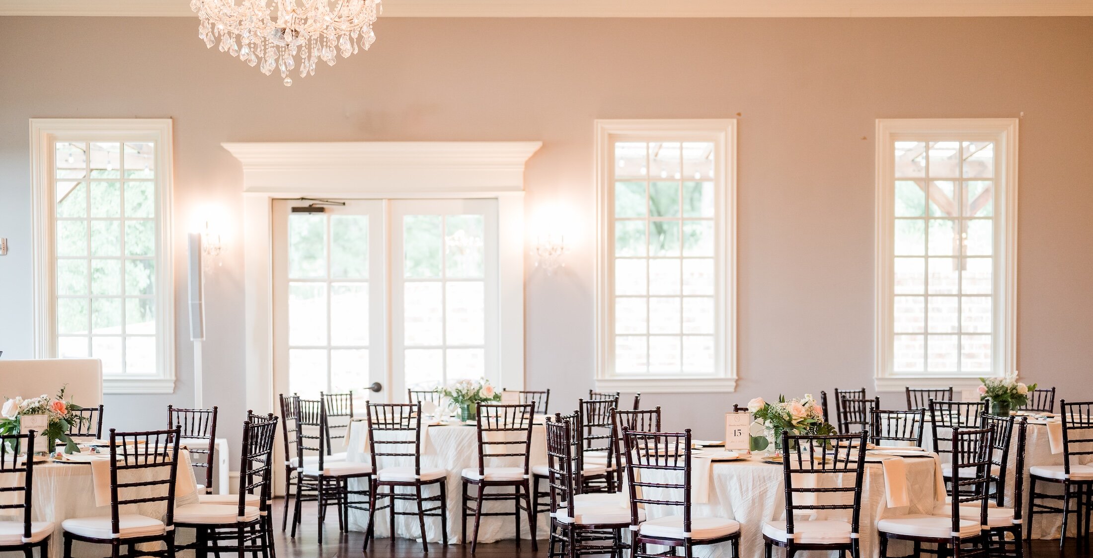 A bright wedding ballroom set for a reception with round tables, dark chiavari chairs, and large windows with white trim.