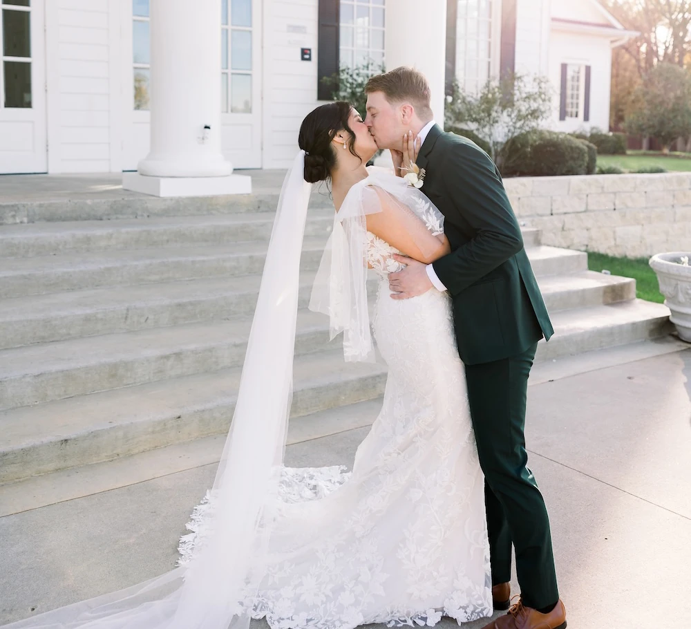 Bride and groom kissing on the steps of a white estate venue with sunlight streaming behind them.