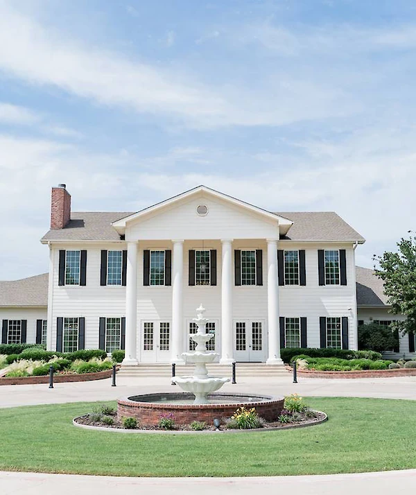 White two-story Southern-style building with tall columns, black shutters, and landscaped shrubs, set against a bright blue sky.