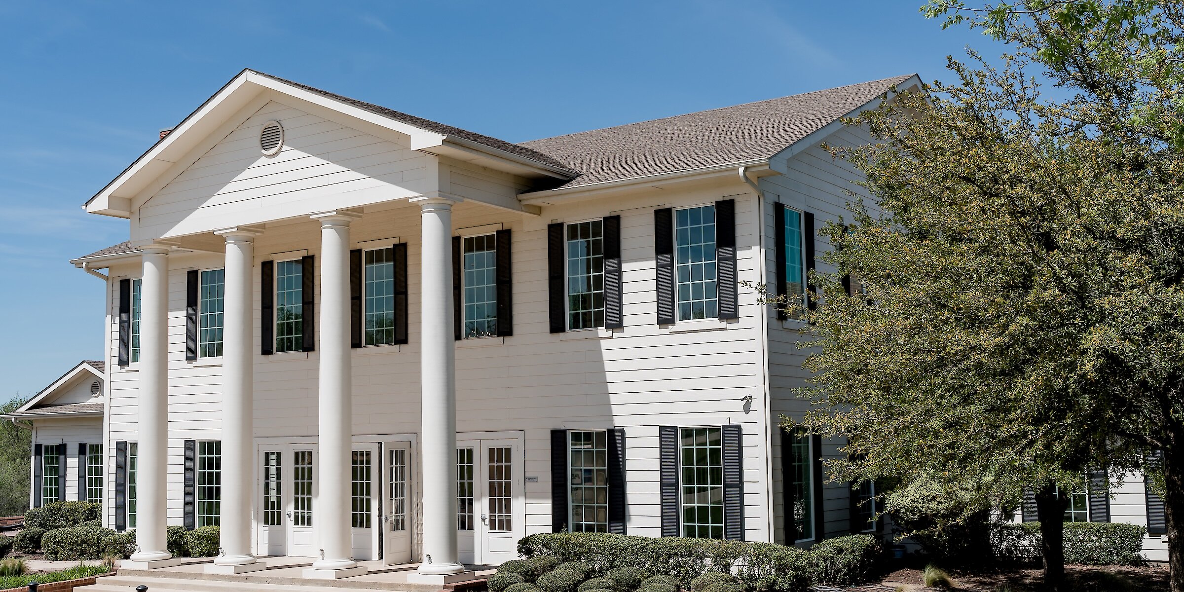 Exterior of a white Southern-style wedding venue with tall columns, black shutters, and manicured landscaping under a clear blue sky.