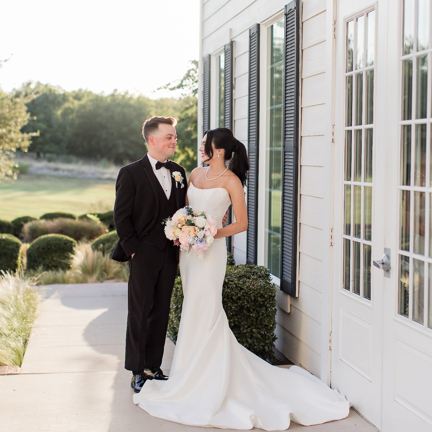 Bride and groom standing outside a white estate venue at sunset, smiling at each other while the bride holds a pastel bouquet.