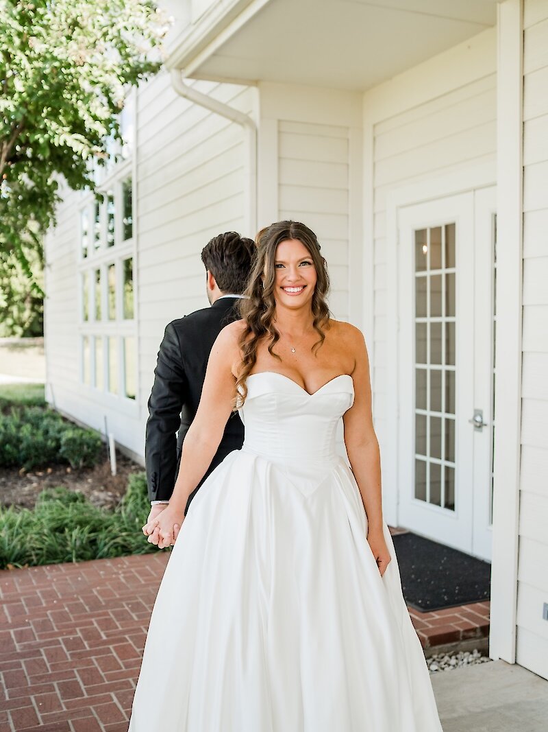 Bride smiling in a strapless ball gown while holding hands with the groom during a first-touch moment outside a white wedding venue.